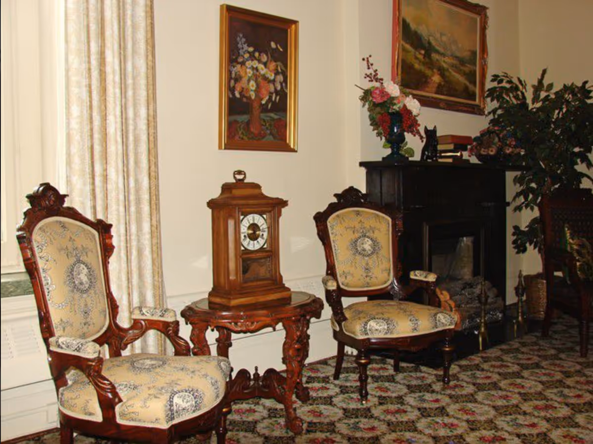 A traditional living room with two ornate wooden armchairs upholstered in patterned fabric, a small wooden side table with a vintage clock on it, a dark wooden fireplace mantel decorated with a vase of flowers, books, and a small black cat figurine. The room has floral carpet, cream-colored curtains, and framed paintings on the walls.