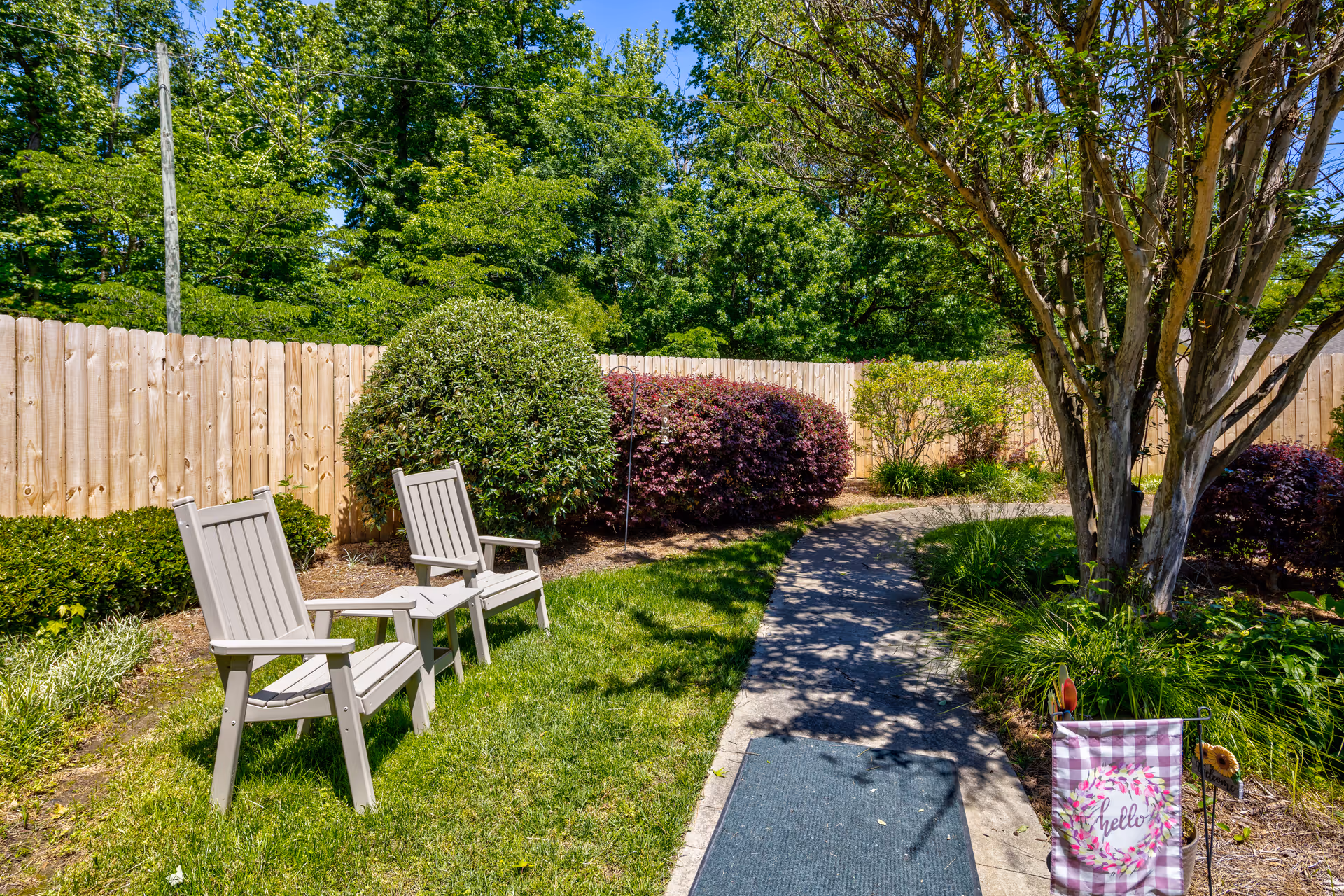 A sunny outdoor garden area with a curved concrete pathway, green grass, two beige wooden chairs with a small matching table between them, surrounded by various bushes and trees, and a wooden fence in the background.