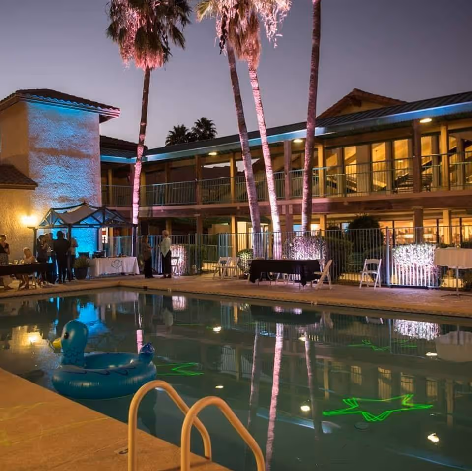 Evening view of an outdoor pool area at a senior living facility with palm trees lit by colorful lights. There are tables and chairs set up around the pool, and a blue inflatable duck float is in the water. The building in the background has large windows and balconies with warm interior lighting.