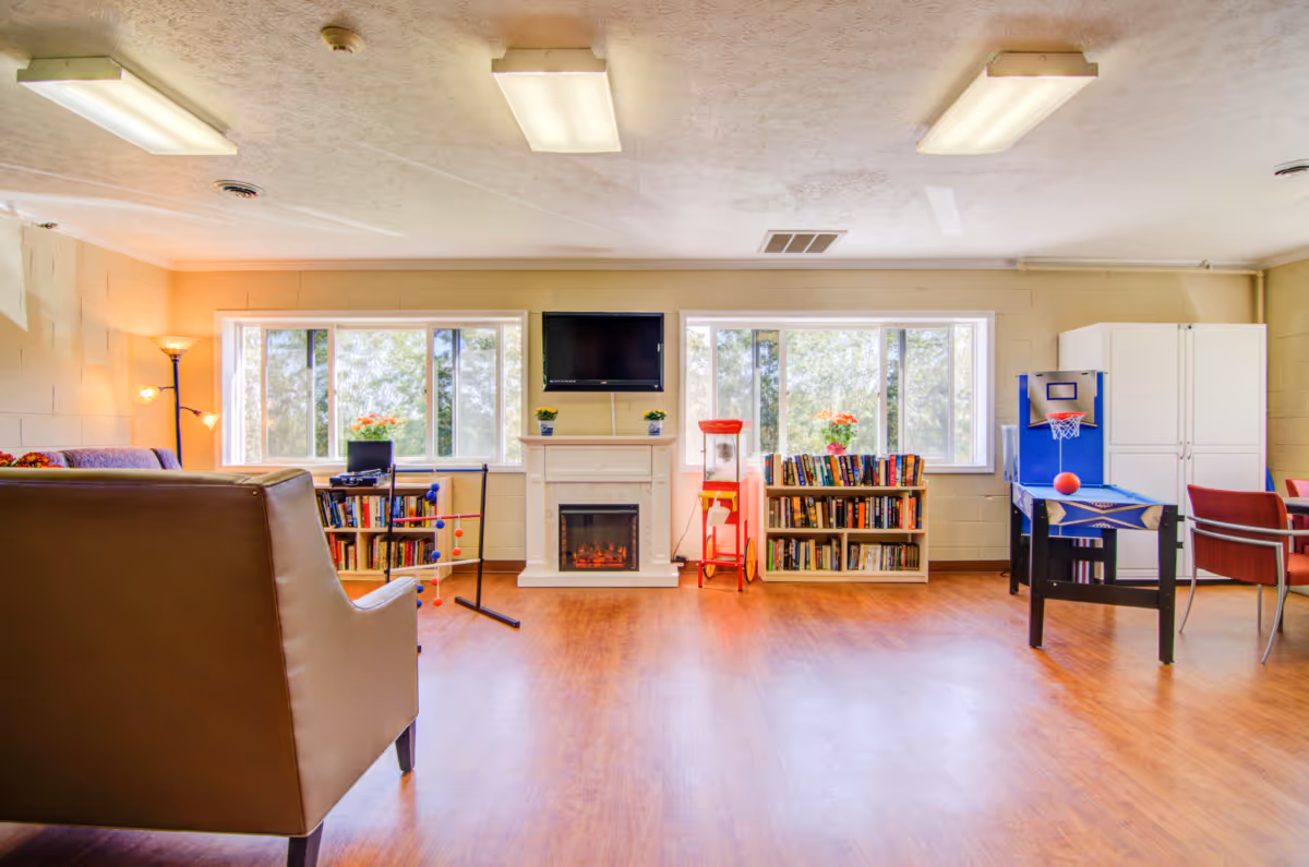 A bright and spacious common area in a senior living facility featuring large windows with views of trees outside. The room has wooden flooring, a white electric fireplace with a TV mounted above it, bookshelves filled with books on either side of the fireplace, a popcorn machine, a small basketball arcade game, and comfortable seating including a leather chair and a sofa. There are also floor lamps providing warm lighting.