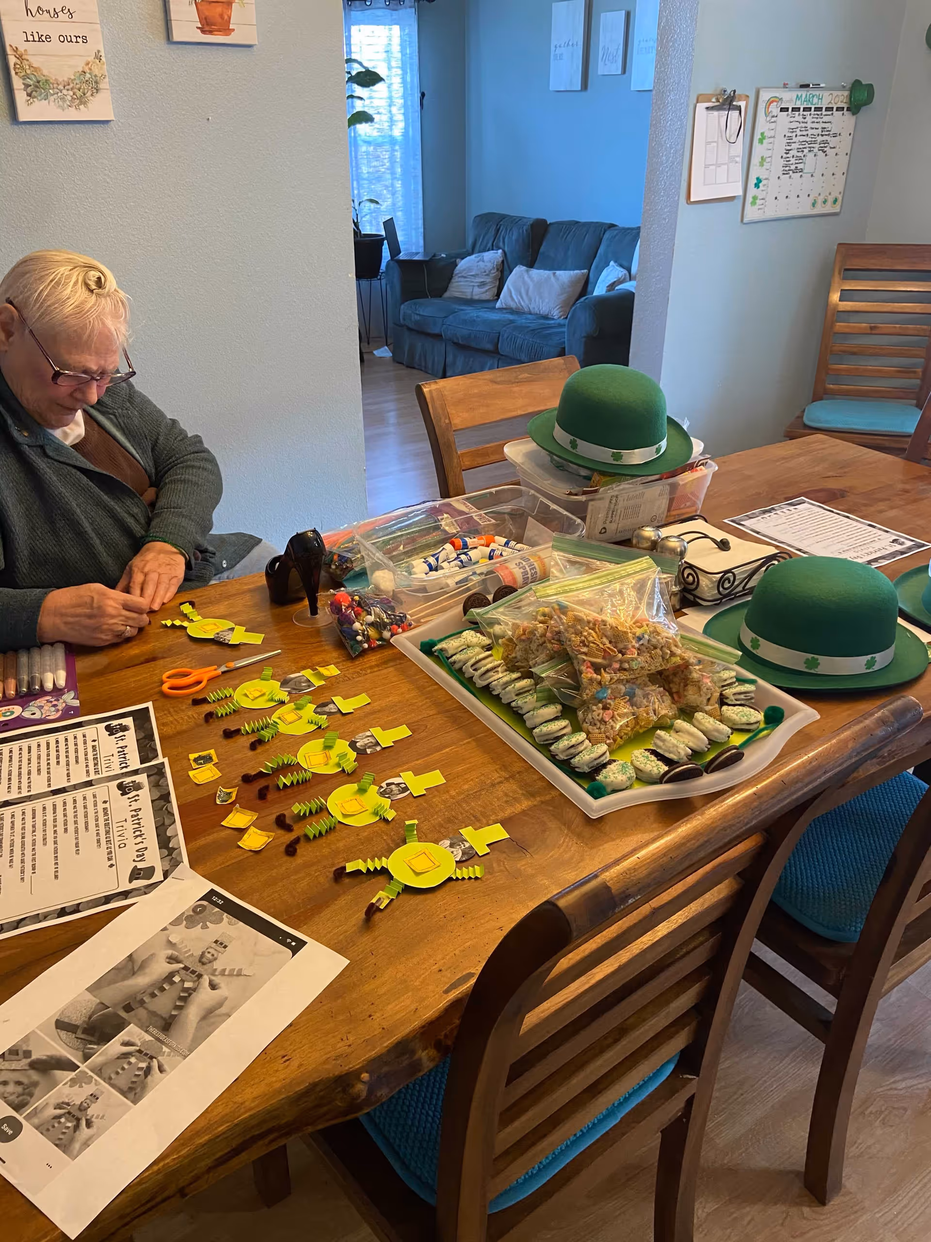 An elderly person sitting at a wooden dining table engaged in a craft activity involving small yellow paper pieces and other craft supplies. The table also has green hats with shamrocks, a tray with decorated cookies and snacks, scissors, and printed instructions. In the background, there is a living room area with a couch and some wall decorations.