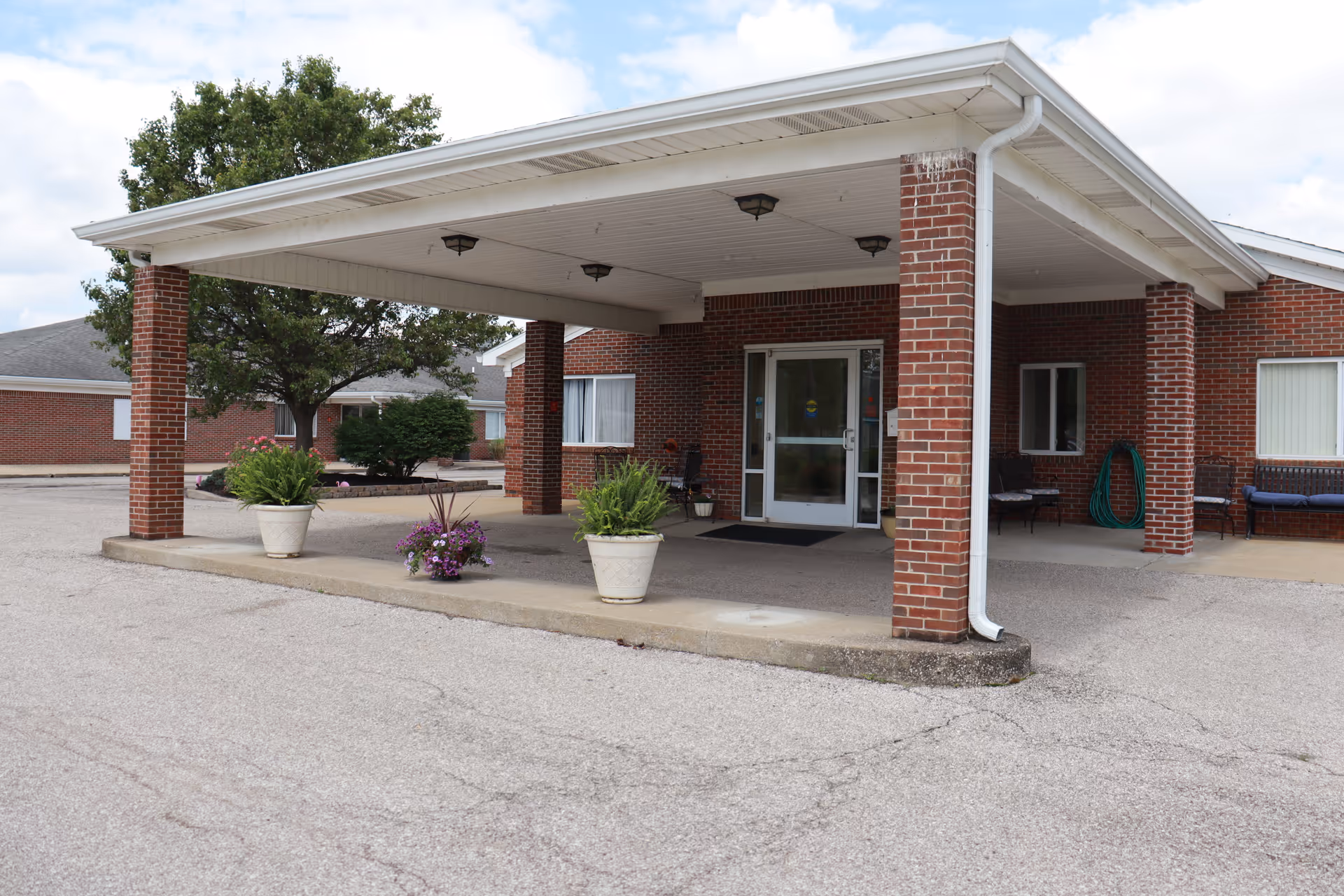 Covered brick porte-cochere entrance with potted plants in front of a single-story senior living building.