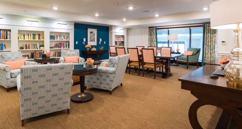 Well-lit communal lounge and dining area with upholstered armchairs, bookshelves, and a long dining table.