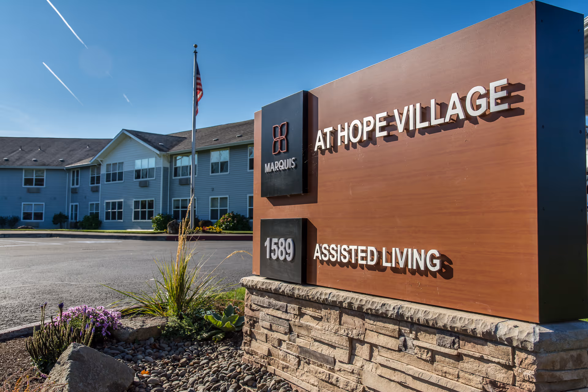 A large wooden sign reading "AT HOPE VILLAGE ASSISTED LIVING" with the number "1589" stands in front of a two-story assisted living building and an American flag.