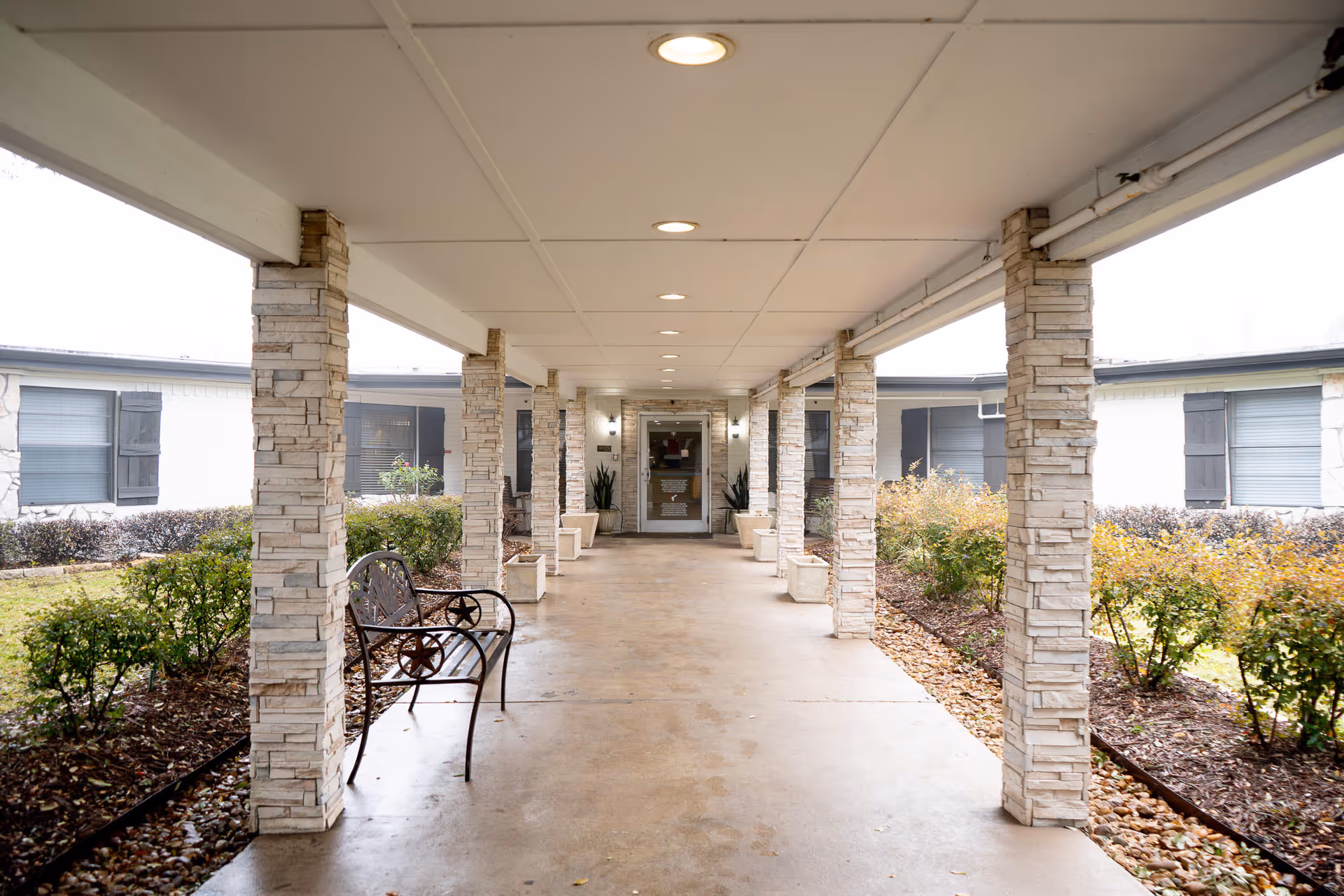 Covered walkway with stone pillars and ceiling lights leading to the entrance of Oak Grove Nursing Home, with benches on the left side and landscaped bushes on both sides.