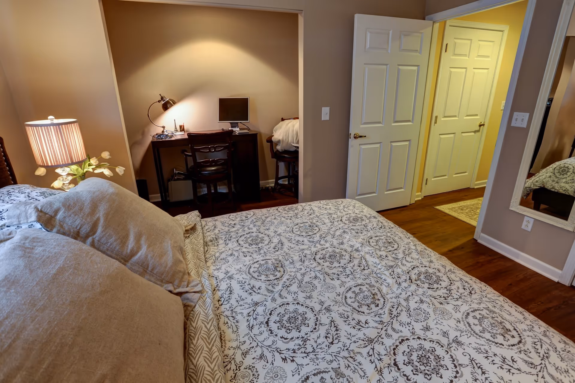 A cozy bedroom with a bed covered in a patterned bedspread and beige pillows. To the left of the bed is a nightstand with a lamp and a small plant. In the background, there is a small workspace with a desk, chair, desk lamp, and a computer monitor. The room has wooden flooring, beige walls, and an open door leading to a hallway with additional doors. A large mirror is mounted on the wall to the right.