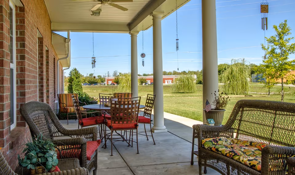 Covered patio with wicker chairs, a dining table with red cushions, hanging wind chimes, and columns overlooking a grassy lawn.