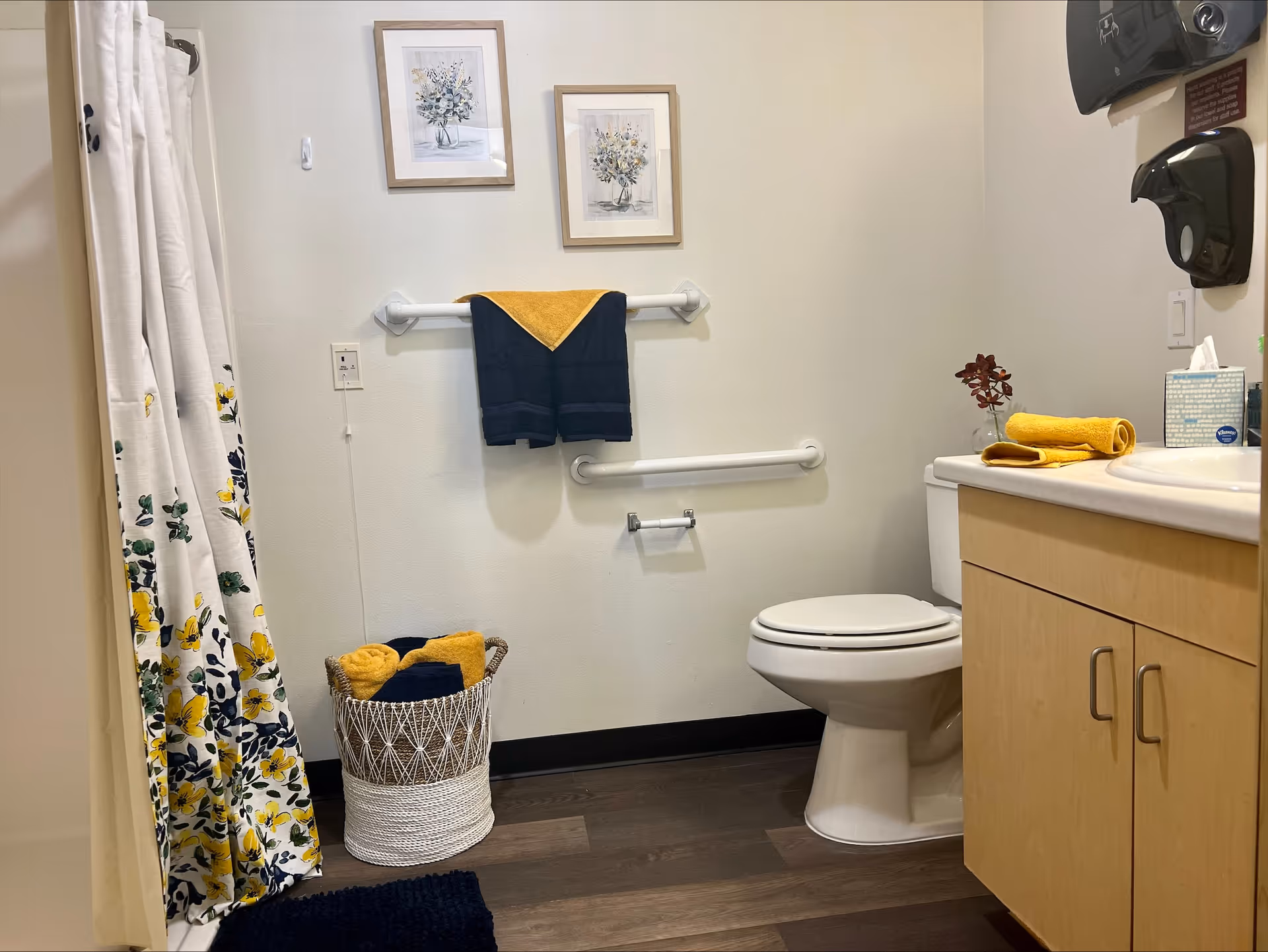 A clean and well-organized bathroom with a toilet, a wooden cabinet with a sink, a shower with a floral curtain, and a basket filled with rolled yellow and navy blue towels. Two framed floral pictures hang on the wall above a towel rack holding a navy blue towel with a yellow corner towel. There are grab bars on the wall near the toilet and a paper towel dispenser mounted above the sink.