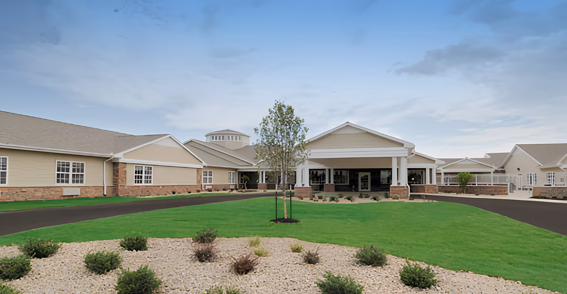 Single-story beige senior living building with a covered entrance, landscaped lawn, and circular driveway under a blue sky.