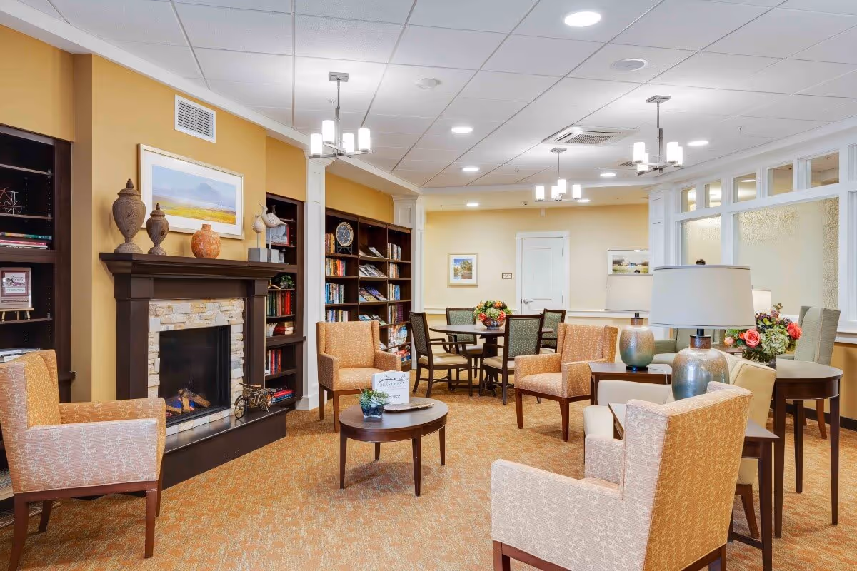 A cozy senior living facility common area with beige and orange armchairs arranged around a small round wooden table. There is a fireplace with a stone surround and a dark wooden mantle, decorated with vases and a framed landscape painting above it. Built-in bookshelves filled with books and decorative items flank the fireplace. In the background, there is a dining area with a round table and chairs, floral centerpieces, and soft overhead lighting from modern chandeliers. The walls are painted in warm yellow and cream tones, and the carpet has a patterned design in complementary colors.