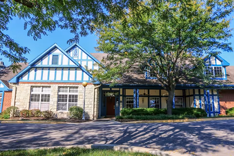 Exterior view of a building with Tudor-style architecture featuring white walls with blue timber framing, large windows, and a brown roof. There are trees and shrubs in front of the building, and a paved driveway or parking area in the foreground under a clear blue sky.