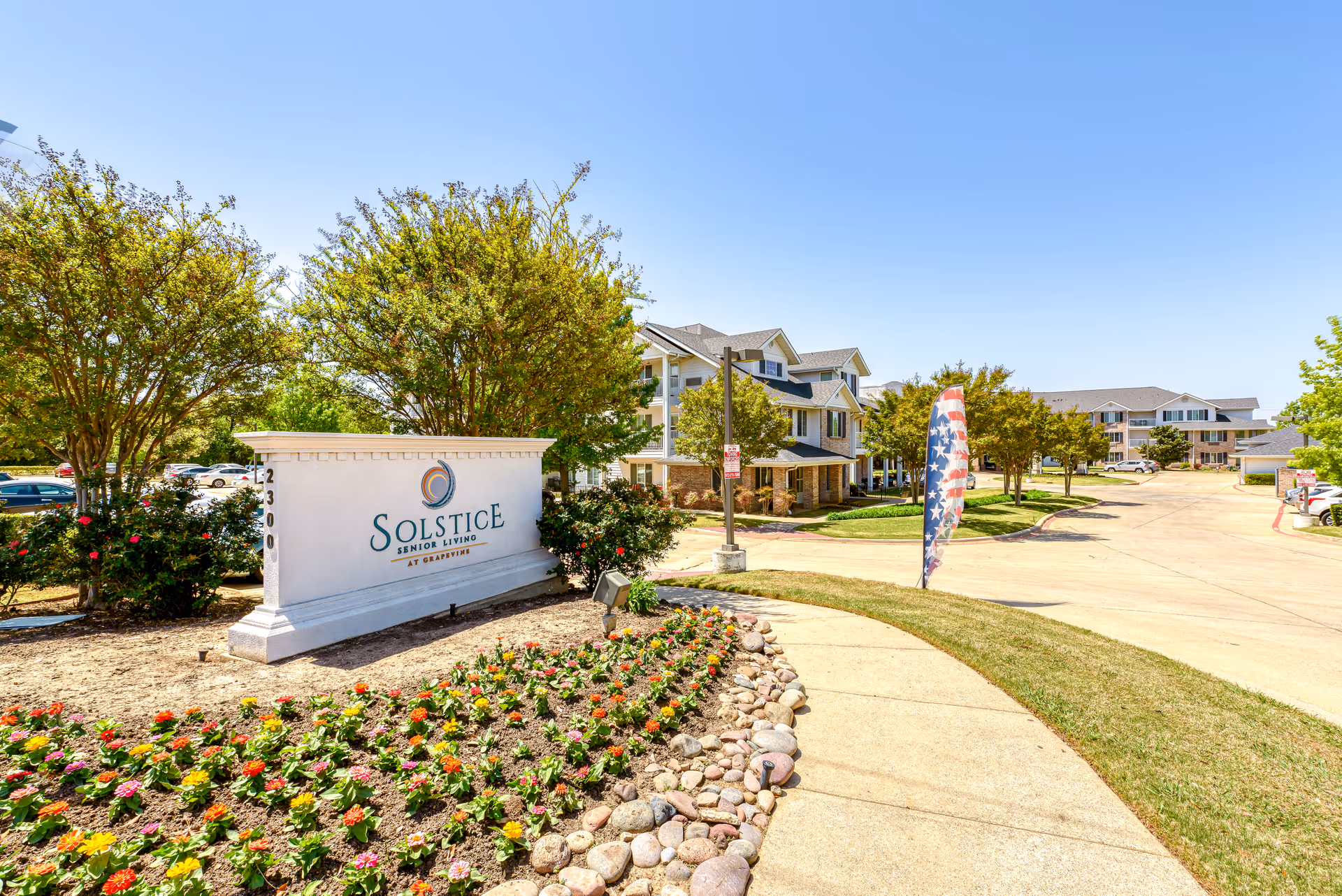 Exterior view of Solstice Senior Living at Grapevine facility entrance with a large sign displaying the facility name, surrounded by landscaped flower beds and trees, with the driveway and buildings visible in the background under a clear blue sky.