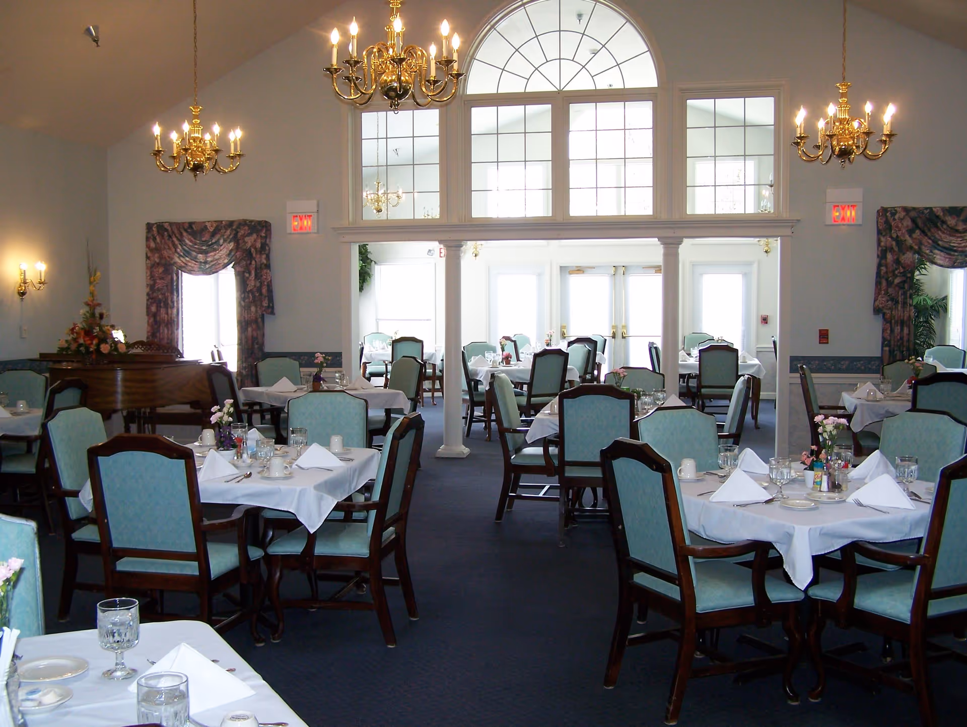 A spacious dining room with multiple tables covered in white tablecloths, each set with glasses, cups, napkins, and small flower arrangements. The room features blue upholstered chairs with wooden frames, blue carpeting, floral curtains, and several brass chandeliers hanging from the ceiling. Large windows and glass doors allow natural light to fill the space.
