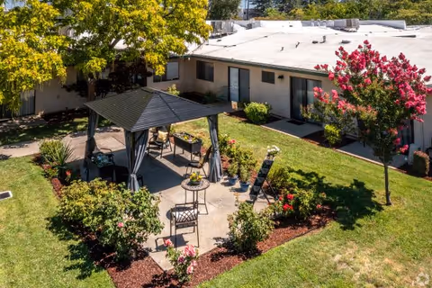 Outdoor courtyard area at Country Club Manor featuring a shaded gazebo with chairs and a small table, surrounded by green grass, flowering bushes, and trees. The building with several doors and windows is visible in the background.