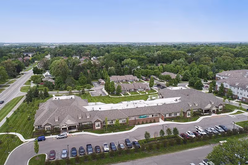 Aerial view of American House Freedom Place Rochester senior living facility showing multiple connected buildings with pitched roofs, surrounded by green lawns, trees, and a parking lot with several cars. Residential houses and dense trees are visible in the background under a clear sky.