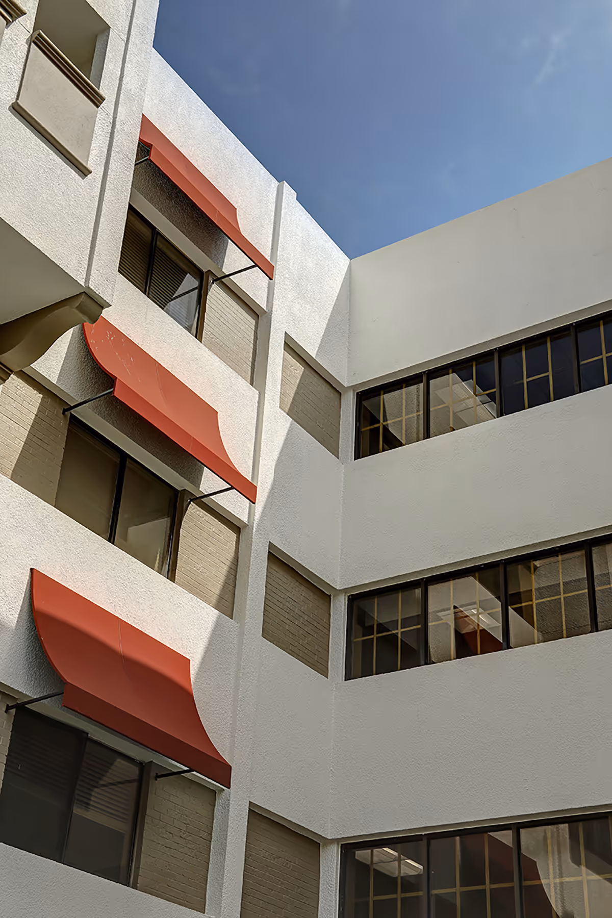 Looking up at the white exterior courtyard walls of a multi-story building with red window awnings and dark-framed windows against a blue sky.
