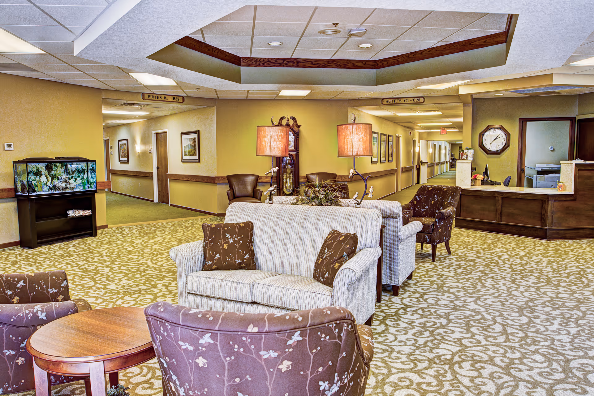 A cozy senior living facility common area with patterned carpet, two beige sofas with brown floral pillows, two brown floral armchairs, a wooden coffee table, and two standing lamps. In the background, there is a hallway with signs indicating suites B1-B32 and C1-C28, a large wall clock, a reception desk, and an aquarium on a stand.