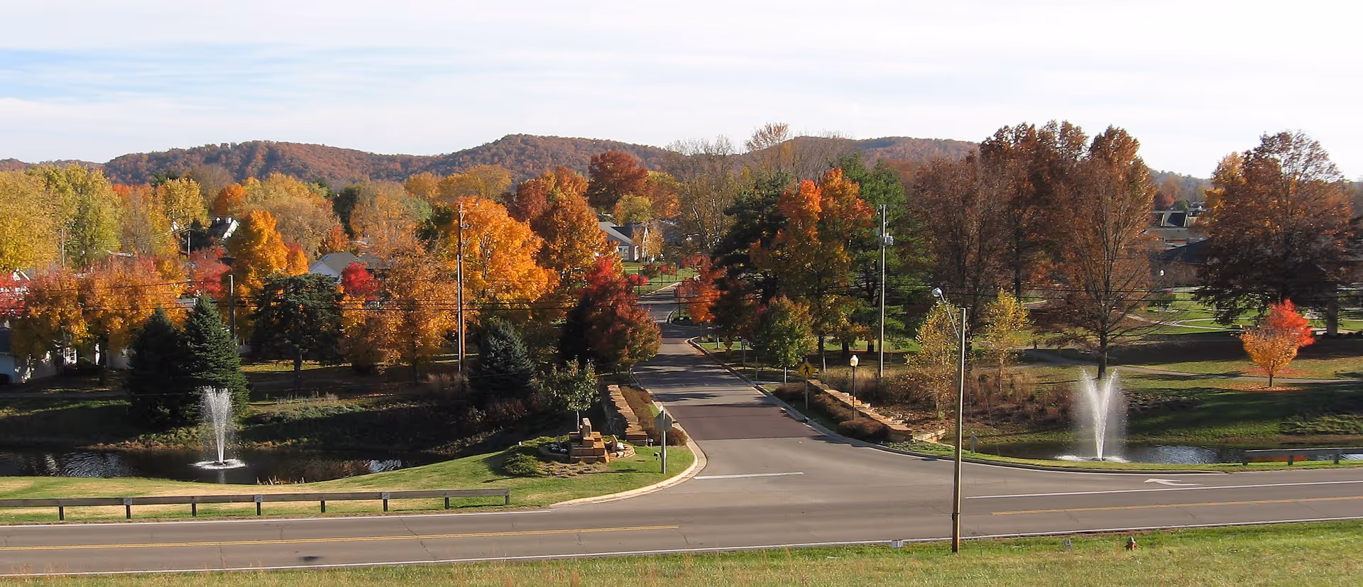 Scenic view of a senior living community entrance with two fountains, a road, and autumn-colored trees and hills in the background.