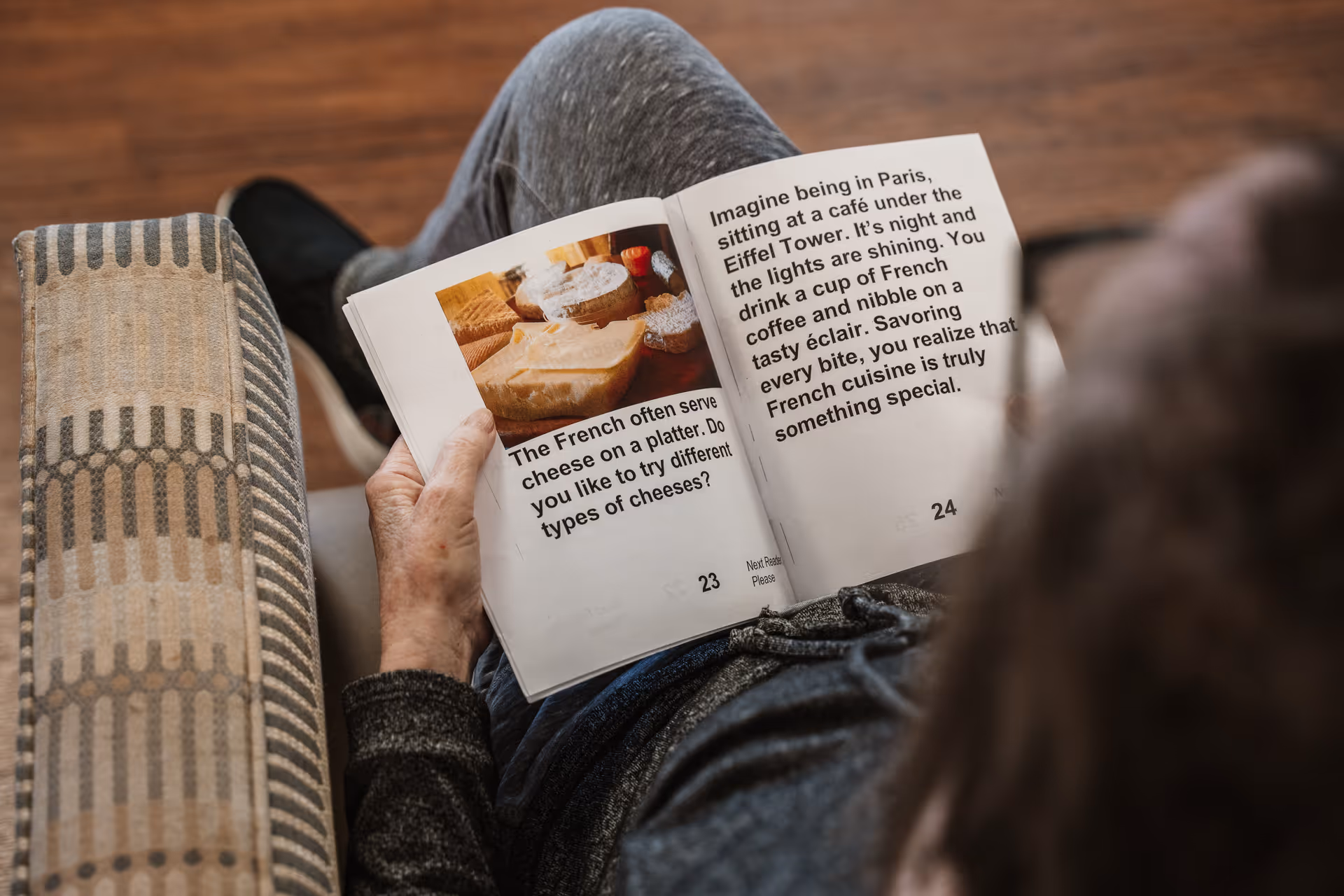 An elderly person sitting on a patterned couch reading a book about French cuisine, showing images and text about French cheese and enjoying coffee and éclairs in Paris.