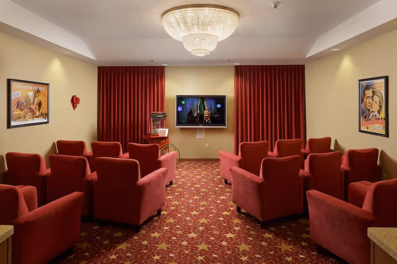 Small theater-style common room with rows of red upholstered armchairs facing a wall-mounted TV, red curtains, patterned star carpet, and vintage movie posters on the walls.