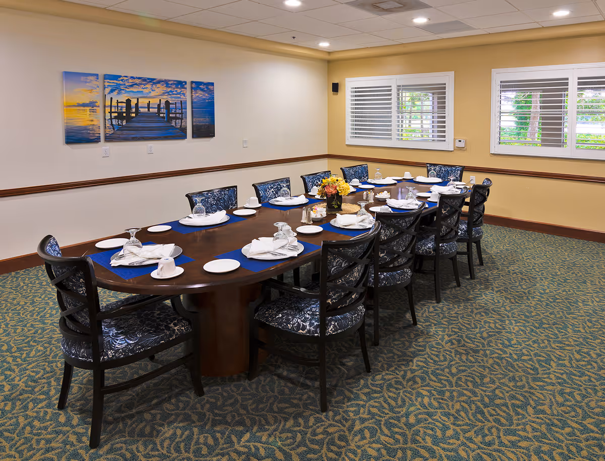 A dining room with a long wooden table set for twelve people. Each place setting includes a white plate, a cup, a napkin, and an upside-down glass. The chairs have dark wooden frames with patterned blue upholstery. The room has beige walls with a wooden chair rail, two windows with white shutters, and a three-panel wall art depicting a pier over water at sunset. The carpet has a blue and green leaf pattern.