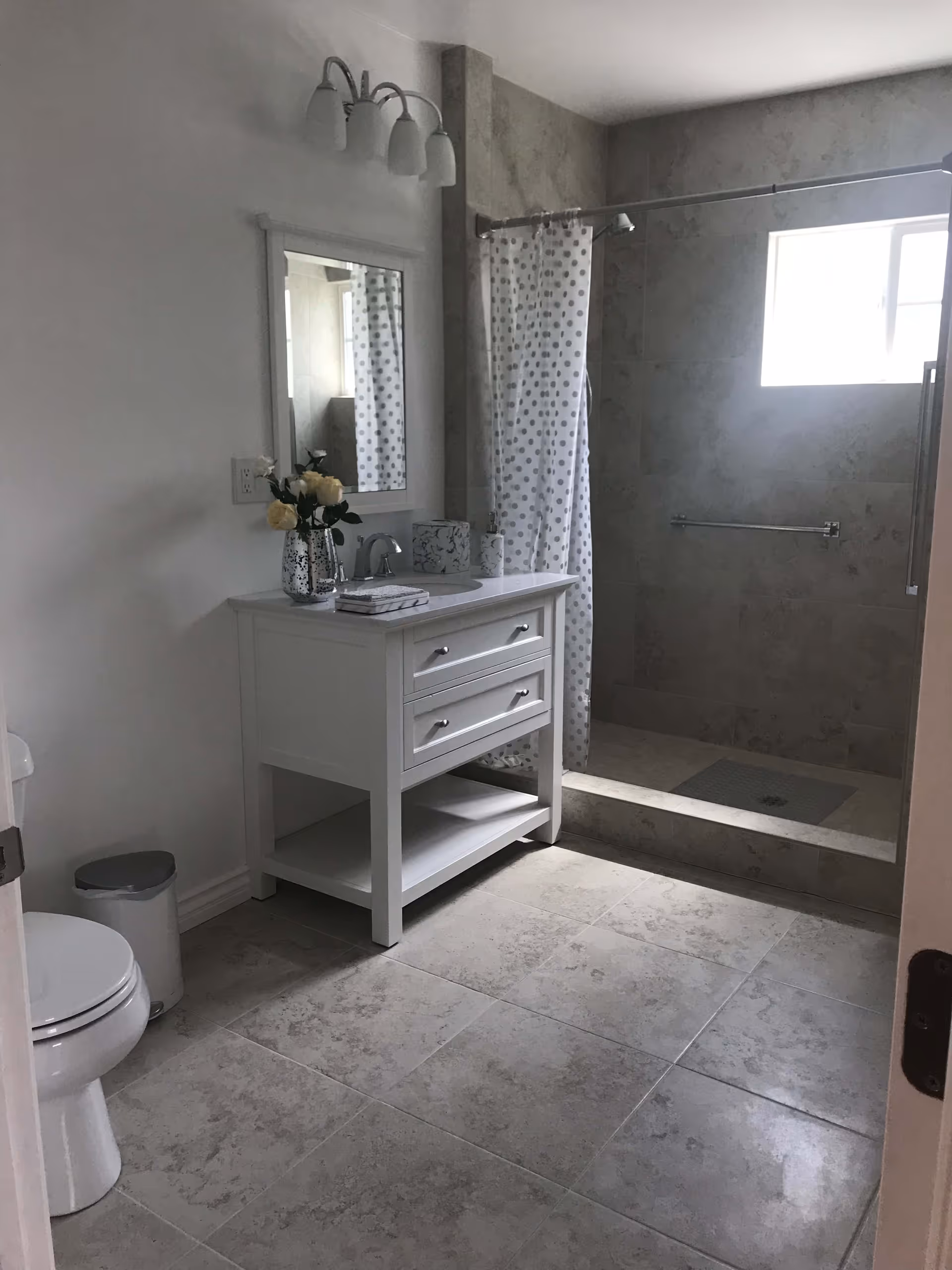 A clean bathroom with a white vanity cabinet topped with a sink and a mirror above it. There is a vase with white flowers on the vanity. To the right is a walk-in shower with a polka dot shower curtain and a small window letting in natural light. On the left side, there is a white toilet and a small trash can. The floor is tiled with large beige tiles.