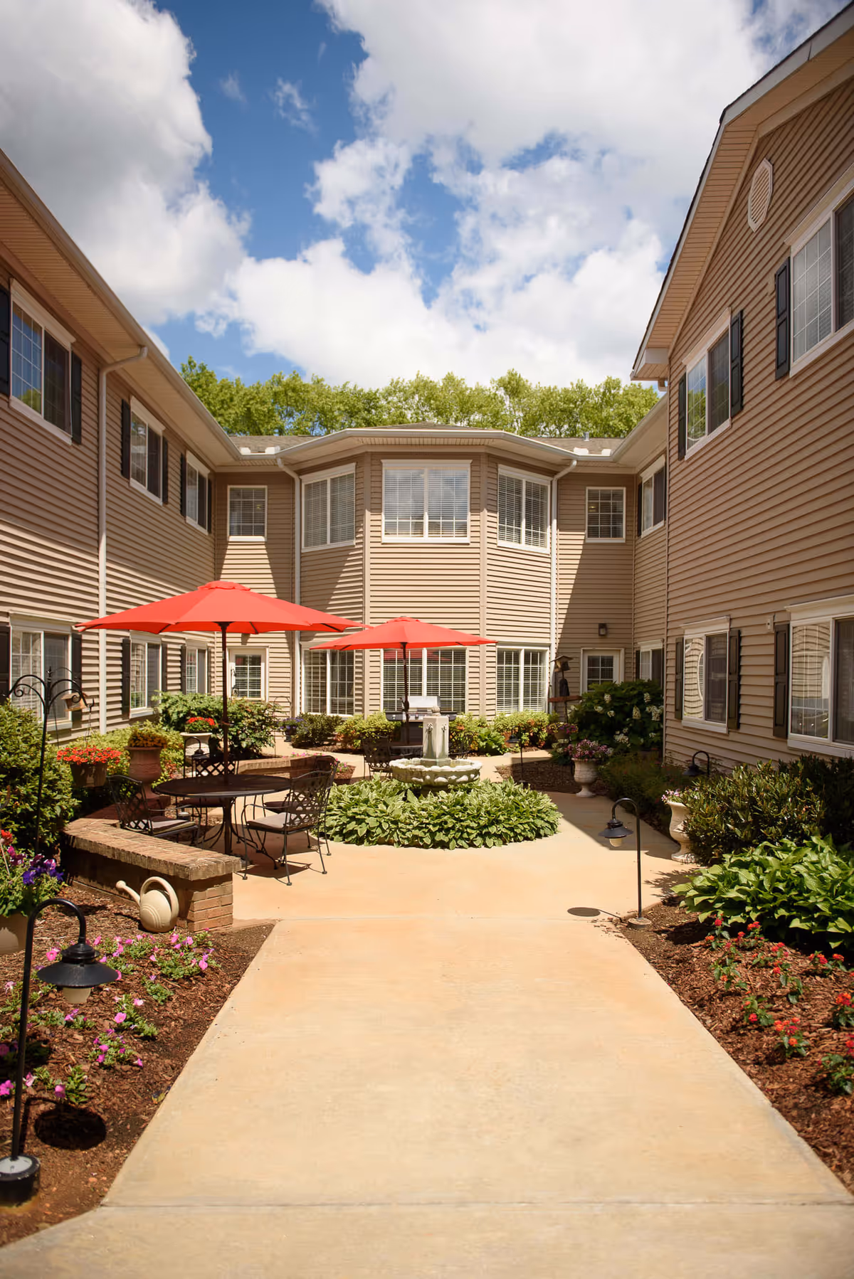 Outdoor courtyard area of a senior living facility with beige siding buildings surrounding it. The courtyard features a concrete walkway, garden beds with flowers and shrubs, a central fountain, and several black metal tables with red umbrellas providing shade.