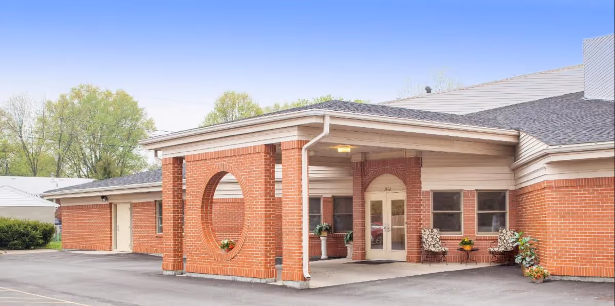 Exterior view of Klondike Nursing and Rehabilitation Center showing a brick building with a covered entrance supported by brick columns. There are two chairs and a small table with plants near the entrance door. Trees and a clear sky are visible in the background.