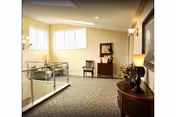 A well-lit interior hallway or common area with beige walls and carpeted floor. There is a glass railing on the left side overlooking a lower level. The area features a wooden side table with a lamp and floral arrangement, a framed picture on the wall, and a wooden chair and small cabinet against the far wall under a window.