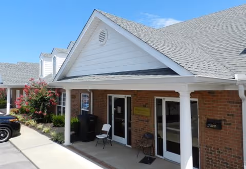 Front entrance of a brick senior living facility with a covered porch, white columns, glass doors, a chair, and a parking area under a clear blue sky.