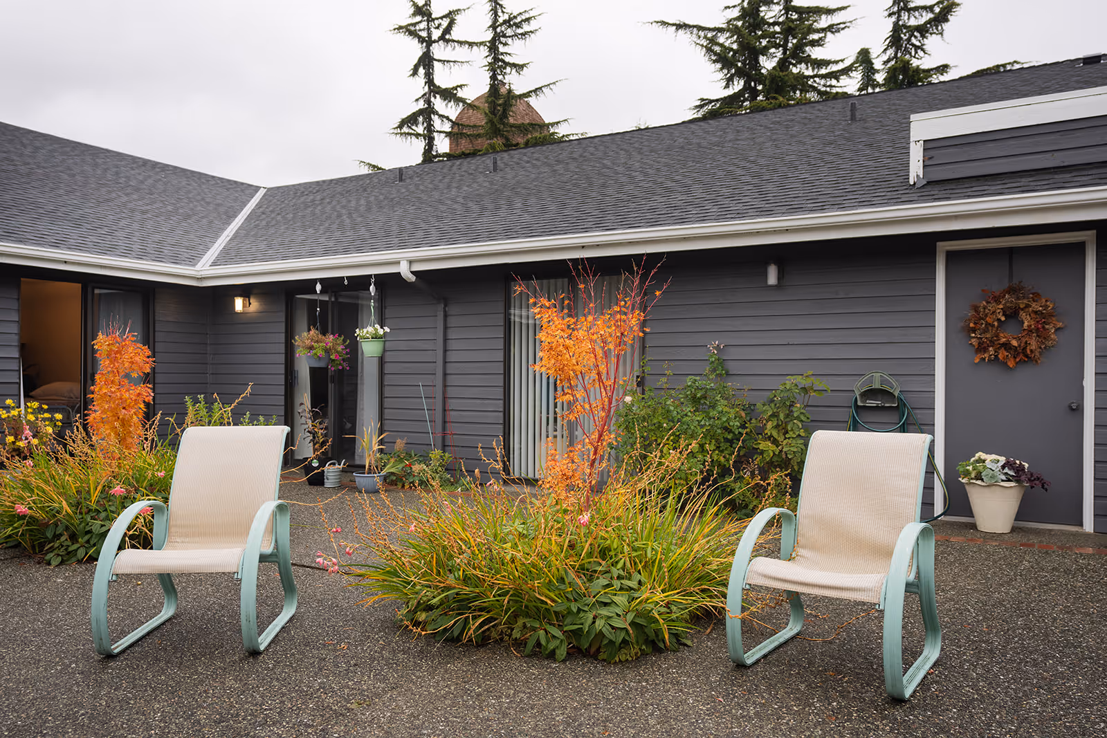 Outdoor courtyard area of a senior living facility with two beige chairs with light blue frames placed on a paved surface. There are plants and shrubs with autumn-colored leaves in the center and along the building. The building exterior is gray with sliding glass doors and a door decorated with a fall wreath. Hanging flower pots are visible near the doors.