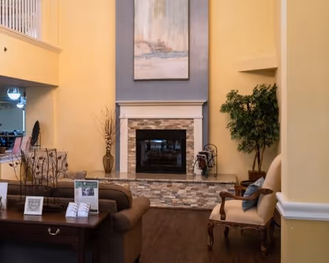 A cozy senior living facility common area with a stone fireplace centered on a blue accent wall. There is a brown sofa and a wooden table with framed photos and decorations in the foreground. To the right, a cushioned armchair with a blue pillow sits next to a potted plant. The walls are painted yellow, and the floor is dark wood.