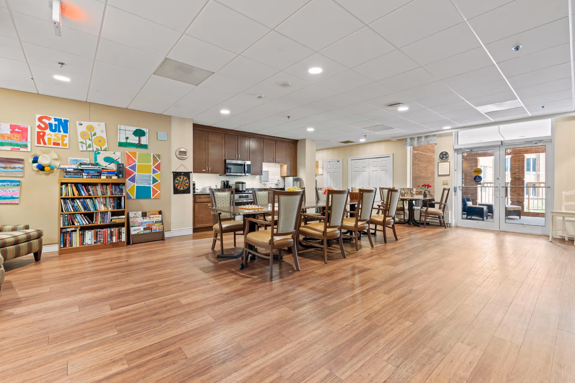 A spacious common area in a senior living facility featuring a dining table with chairs, a kitchenette with wooden cabinets and appliances, a bookshelf filled with books, colorful artwork on the wall, and large glass doors leading to an outdoor seating area.