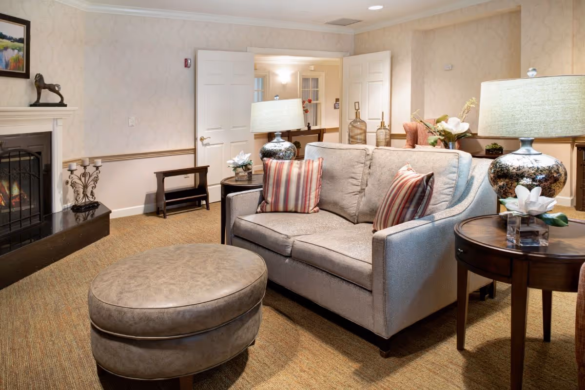 A cozy living room area with a beige loveseat adorned with two striped pillows, a round leather ottoman, two side tables each with a decorative lamp and flower arrangement, a fireplace with a metal screen, and a small bench against the wall. The room has beige carpet, light wallpaper, and double doors leading to another room.