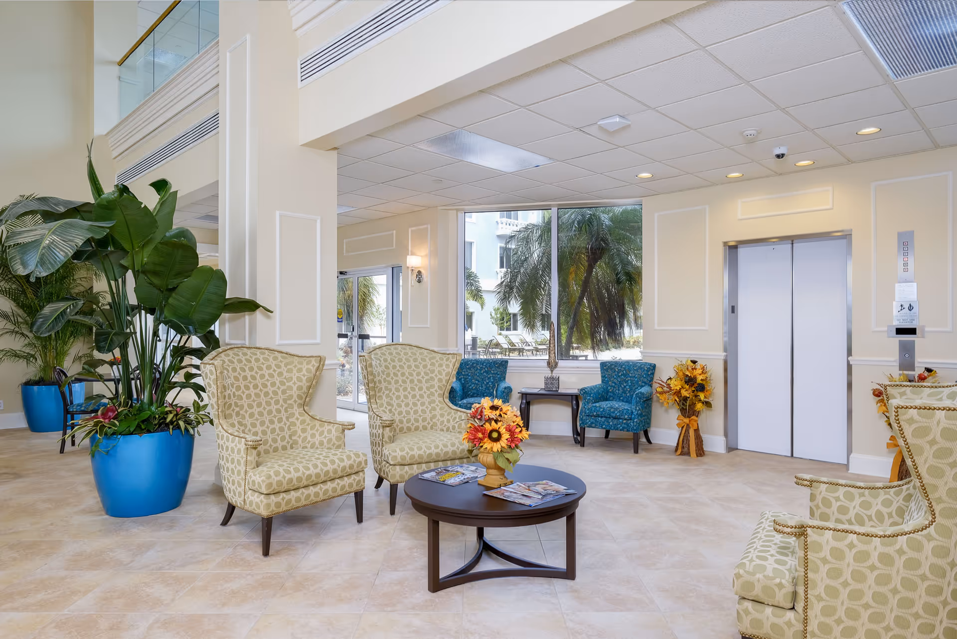 A bright and spacious seating area in a senior living facility with patterned armchairs arranged around a round wooden coffee table holding a floral arrangement and magazines. Large windows reveal palm trees outside, and there is an elevator with closed doors on the right side. The room features beige tiled floors, cream-colored walls, and large potted plants in blue pots.