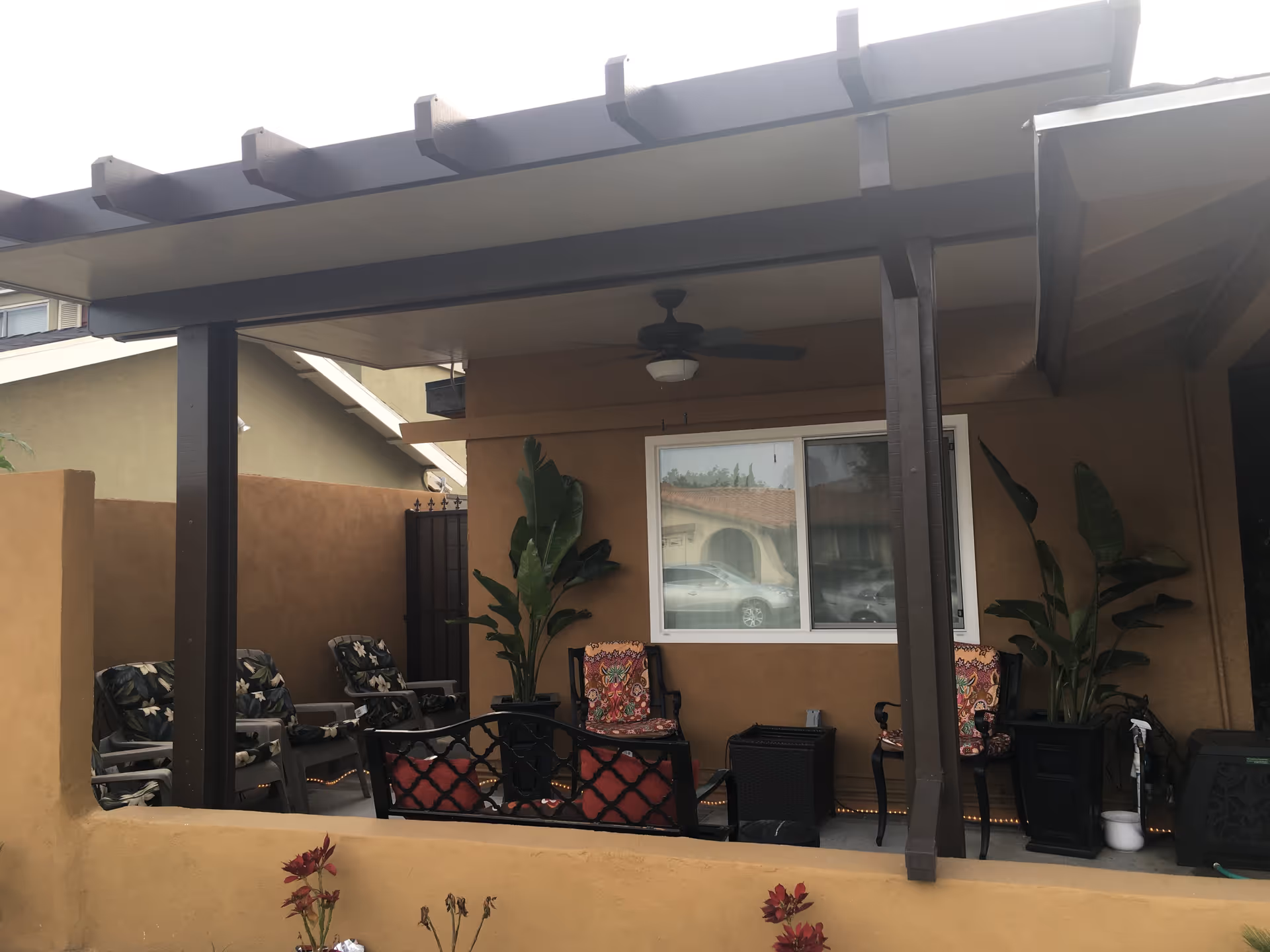 Covered outdoor patio area with several cushioned chairs, potted plants, a ceiling fan, and a window reflecting a car and neighboring building.