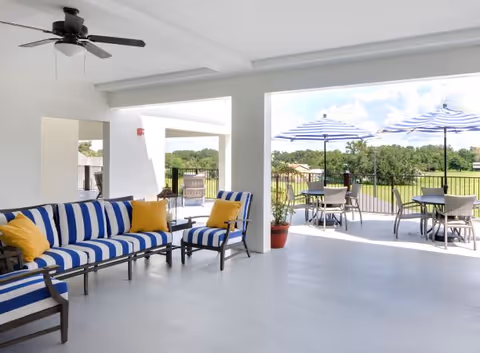 Covered patio area with blue and white striped cushioned seating and yellow pillows, ceiling fan, and open view to an outdoor space with tables, chairs, and blue and white striped umbrellas overlooking a green landscape.
