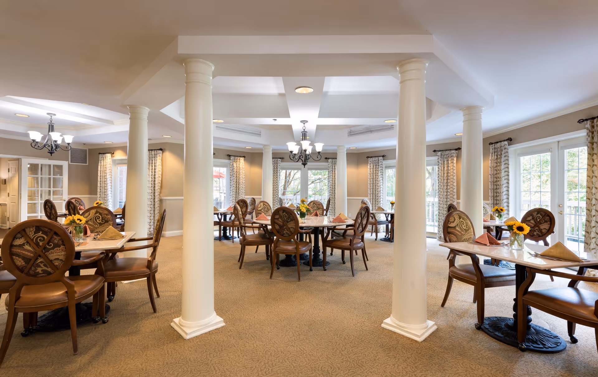 A spacious dining room in a senior living facility with multiple round and rectangular tables surrounded by wooden chairs with patterned upholstery. Each table is set with folded napkins and small vases containing sunflowers. The room features beige carpeting, cream-colored walls, large windows with patterned curtains, and white columns supporting the ceiling. Chandeliers provide lighting, and natural light streams in through the windows.