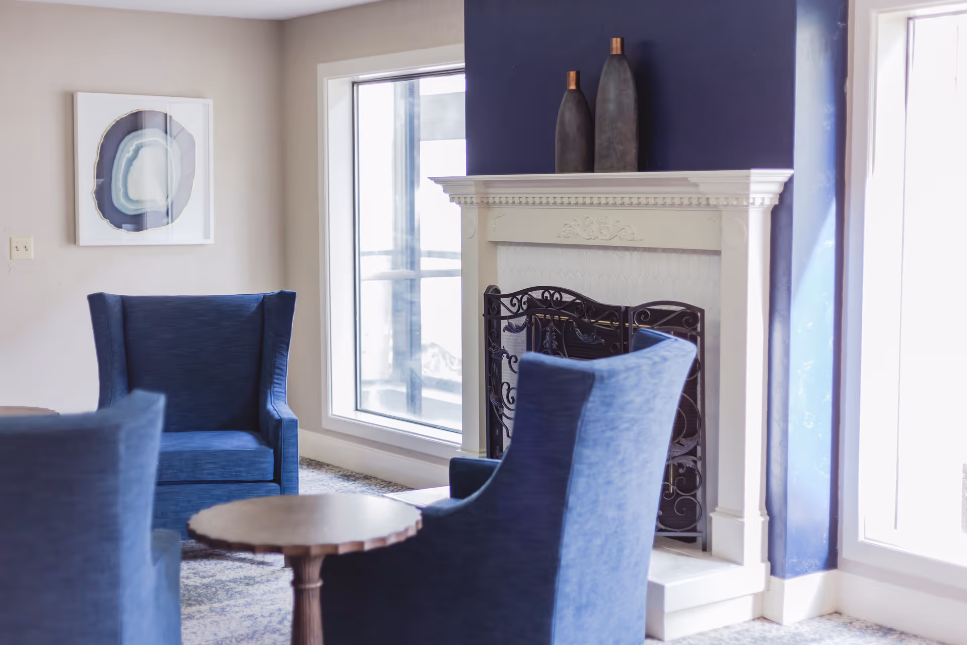 A cozy seating area with three blue upholstered armchairs arranged around a small wooden round table. Behind the chairs is a white fireplace with a decorative black metal screen and two tall vases on the mantel. The wall behind the fireplace is painted dark blue, and there are large windows letting in natural light.