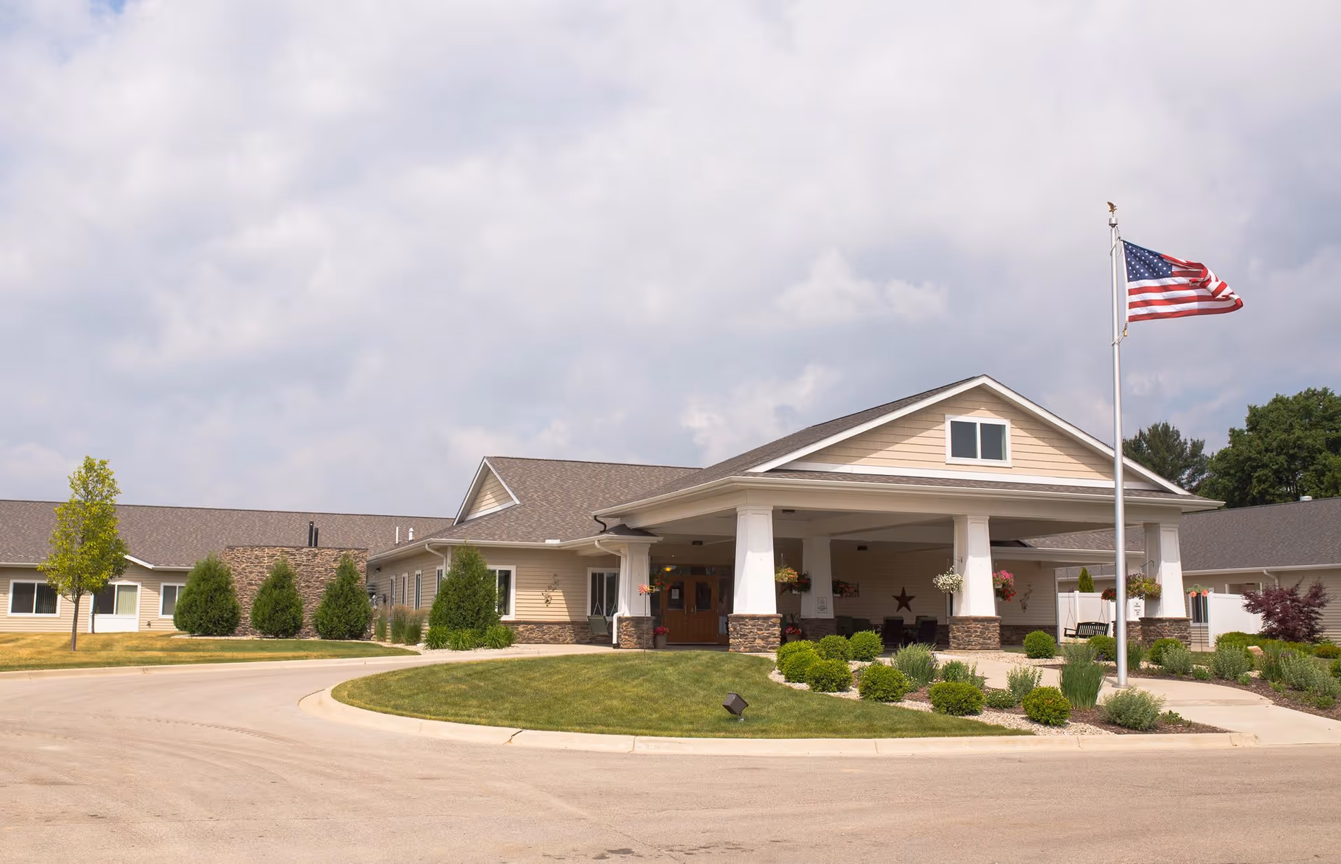 Exterior view of Covenant Glen of Frankenmuth facility showing a single-story building with a covered entrance, an American flag on a flagpole, landscaped bushes, and a partly cloudy sky.