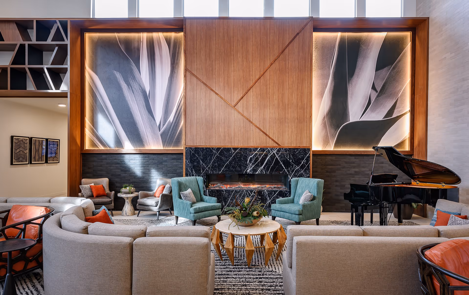 A modern and stylish living room area with a large sectional sofa, two teal armchairs, and a round wooden coffee table with a plant centerpiece. The room features a black marble fireplace with a wooden panel above it, flanked by two large illuminated abstract art pieces. A black grand piano is positioned on the right side of the room. The space is decorated with accent pillows in orange and gray tones and has a patterned rug on the floor.