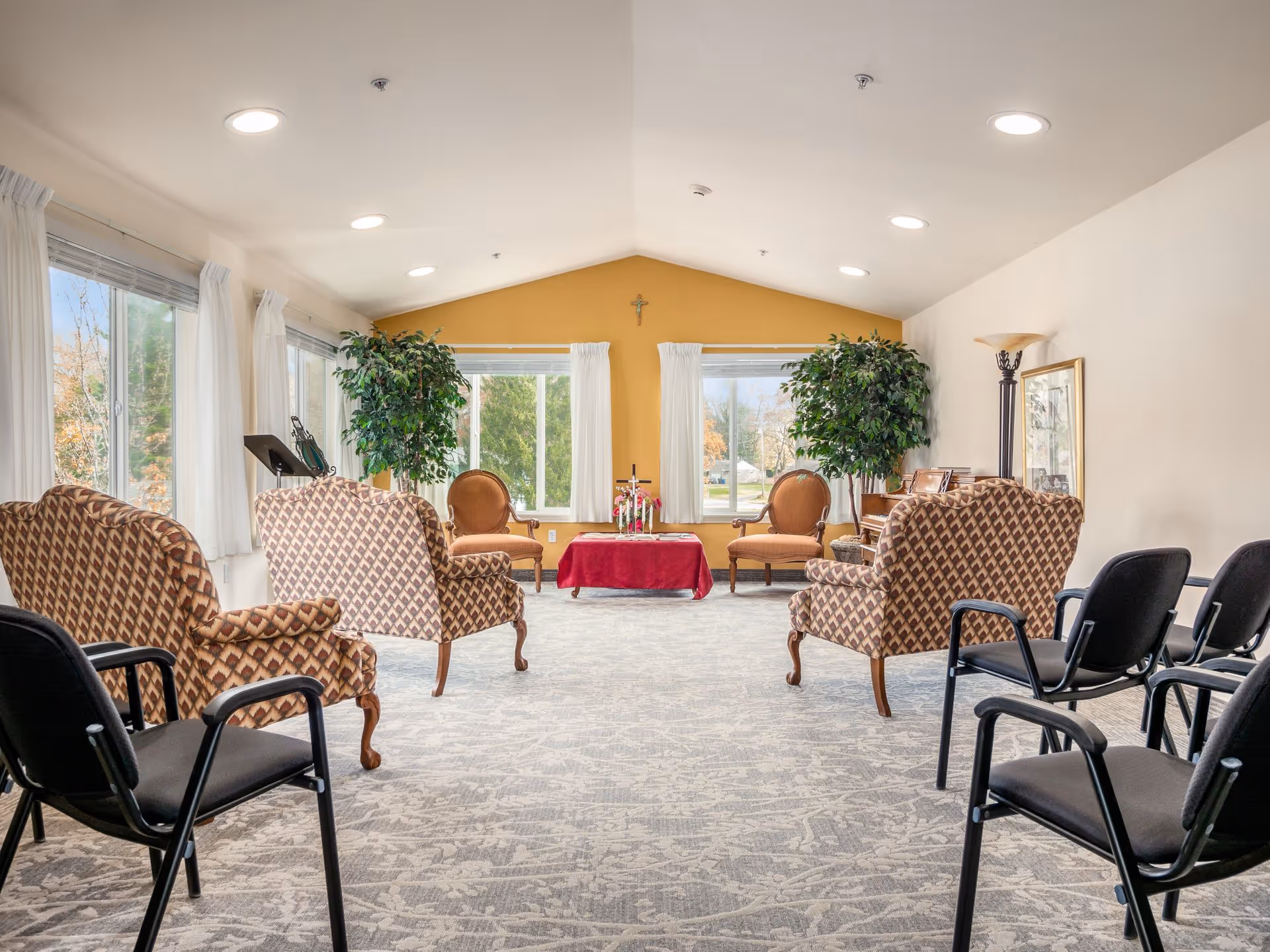 A bright, spacious room with large windows and white curtains letting in natural light. The room features patterned armchairs and black chairs arranged in a semi-circle facing a small table covered with a red cloth. Two large potted plants flank the windows, and a cross is mounted on the yellow accent wall at the far end. The carpet has a subtle floral pattern, and a floor lamp and framed artwork are visible on the right side.