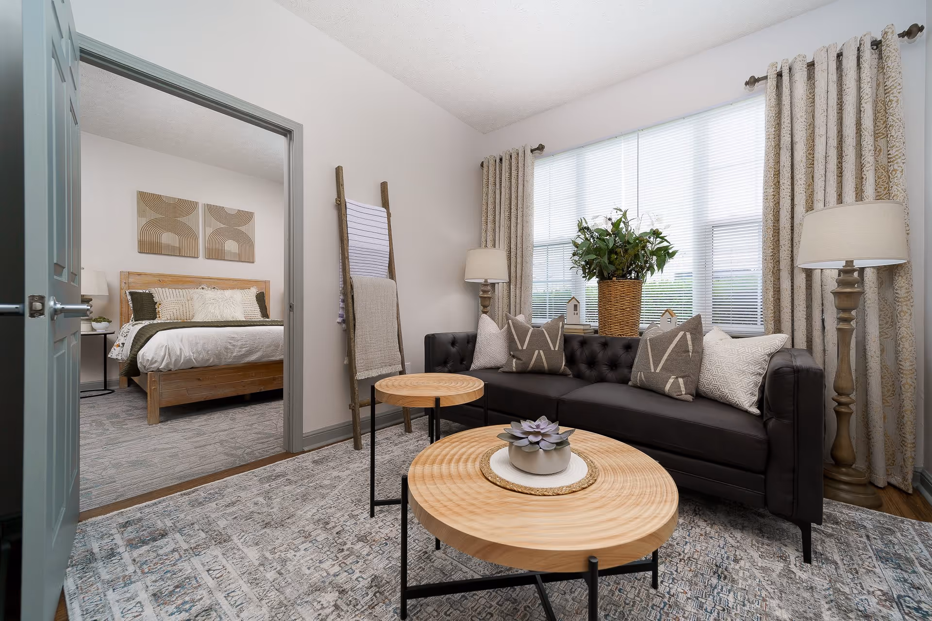 A cozy living room with a black tufted sofa adorned with patterned throw pillows, two round wooden coffee tables with a small plant centerpiece, a tall floor lamp, and a large window with beige curtains. Adjacent to the living room is a bedroom with a wooden bed frame, white bedding, decorative pillows, and abstract wall art above the bed.