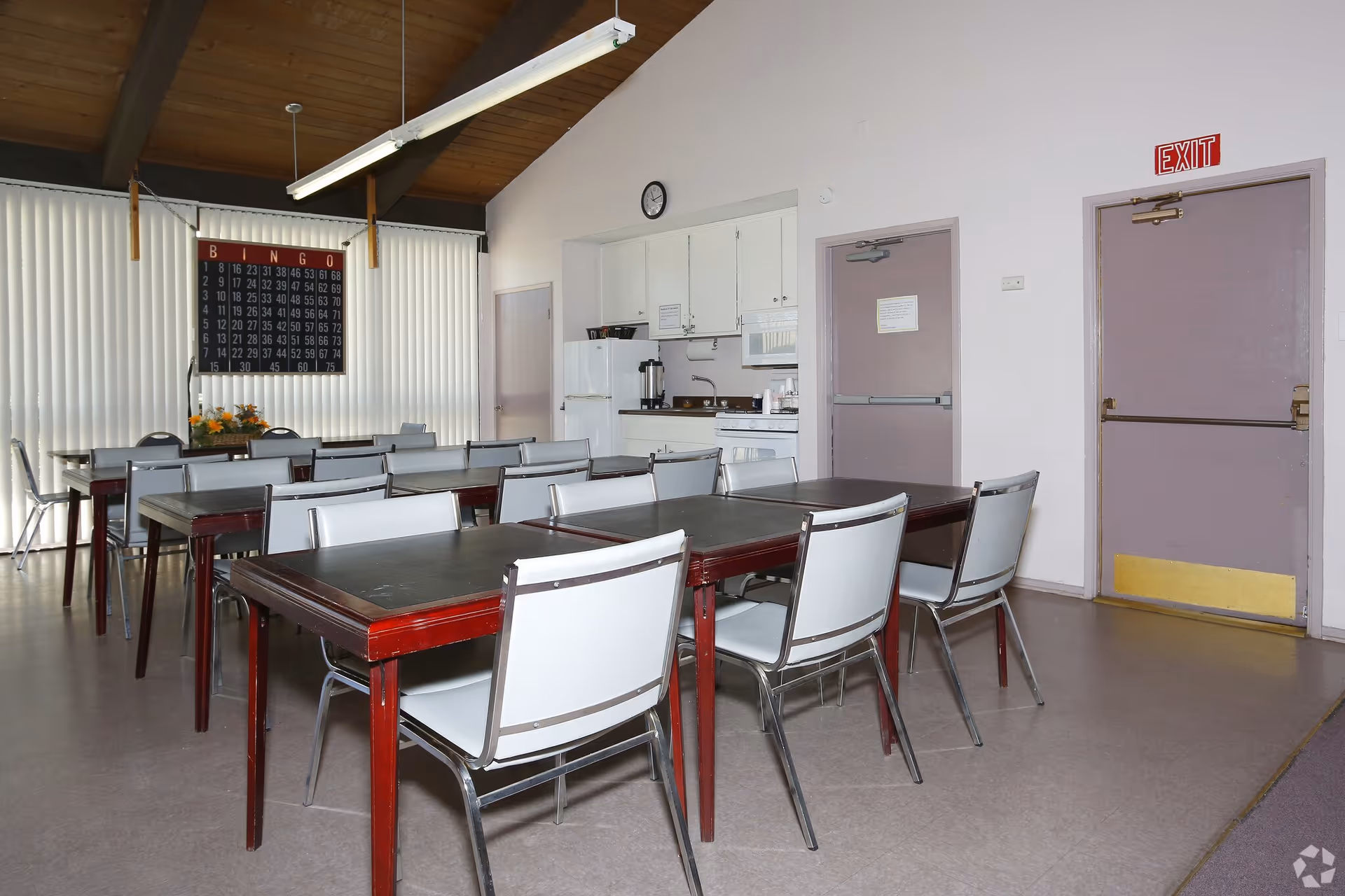 A communal room with several tables and chairs arranged in rows. The room has a high wooden ceiling with exposed beams and a fluorescent light fixture. On the back wall, there is a large bingo board and vertical blinds covering the windows. To the right, there is a small kitchenette area with white cabinets, a refrigerator, a stove, and a coffee maker. Two purple exit doors are visible on the right side of the room.
