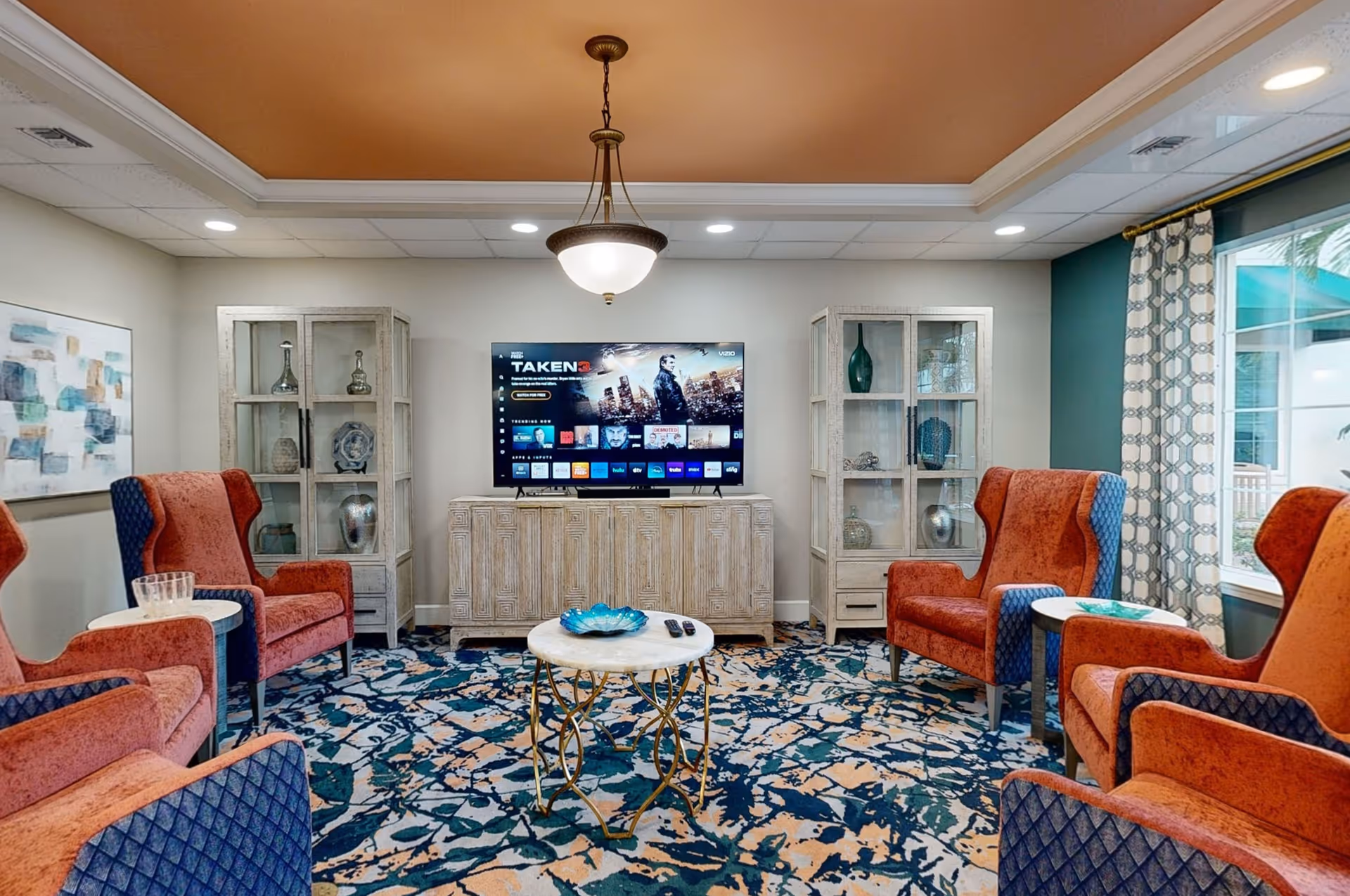 A cozy living room with four orange and blue armchairs arranged around a small round table with a decorative bowl and remote controls. A large flat-screen TV is mounted on a wooden cabinet against the wall, flanked by two glass display cabinets with decorative items. The room has a patterned carpet, a ceiling light fixture, and a large window with patterned curtains letting in natural light.