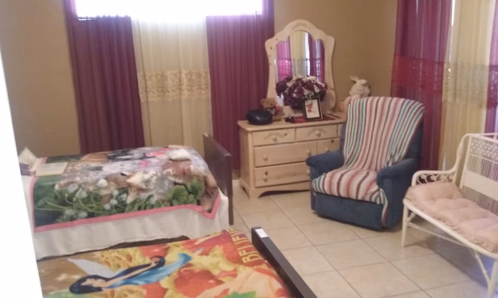 A cozy bedroom with two beds covered in colorful blankets, a wooden dresser with a mirror, a blue armchair with a striped cover, a white cushioned bench, and purple and cream curtains on the windows.