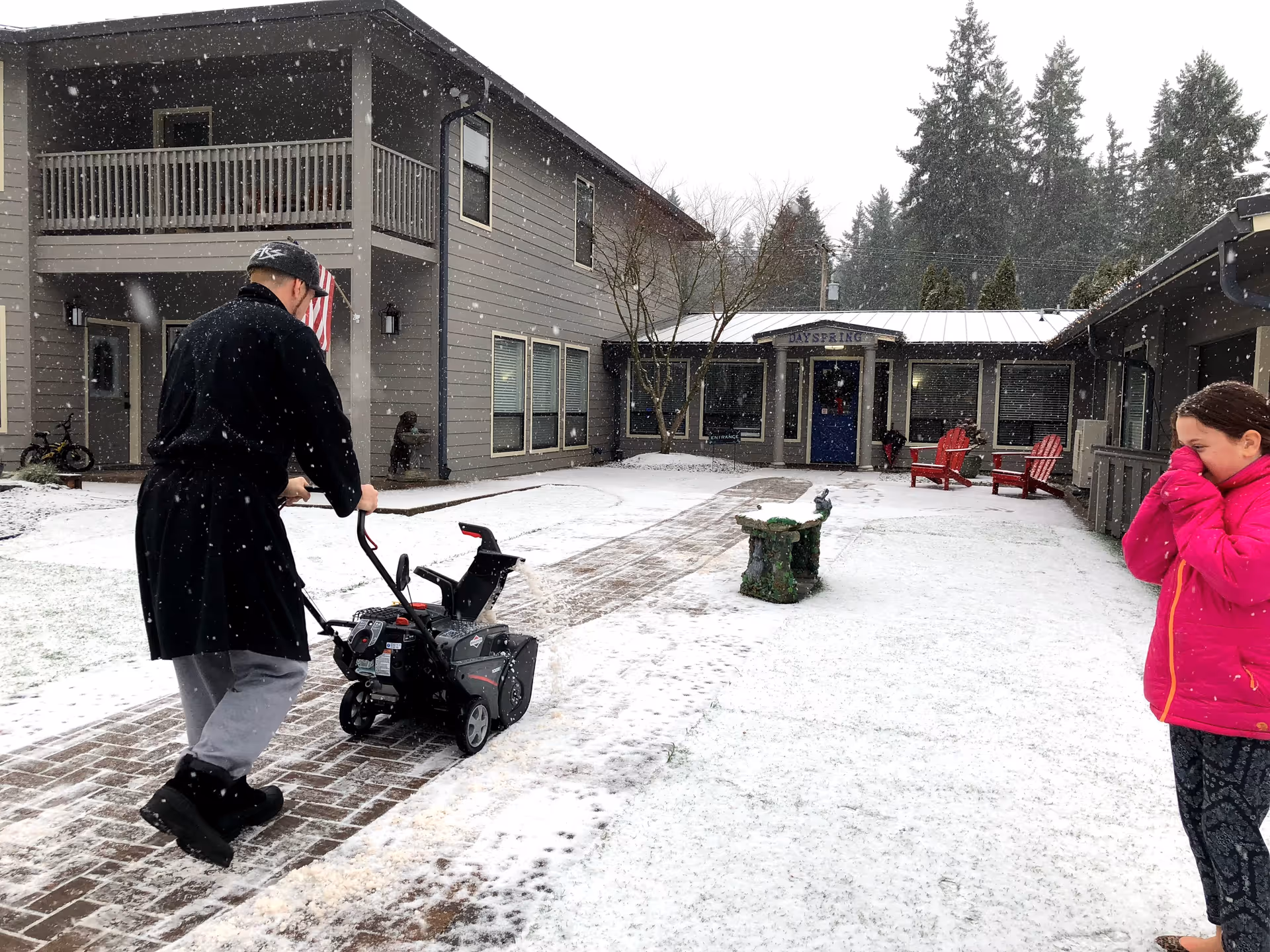 A person wearing a black coat and hat is using a snow blower on a brick pathway in front of a gray building with a sign that reads 'Dayspring'. Snow is falling and covering the ground. A child in a bright pink jacket stands to the right, covering their face from the cold. There are red chairs and some trees in the courtyard area.