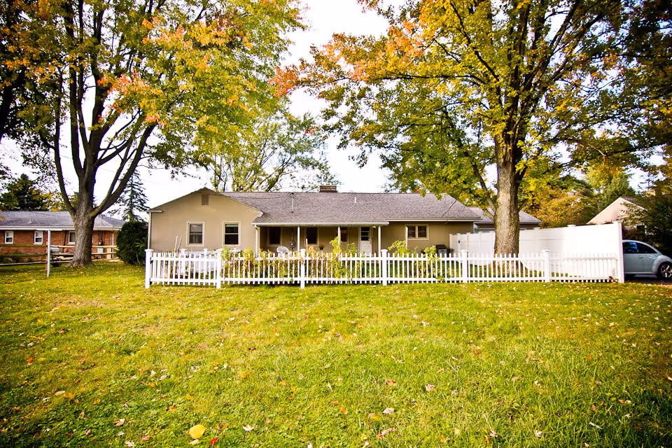 Single-story beige house with a gray roof surrounded by a white picket fence. The house is set in a grassy yard with two large trees displaying green and yellow leaves, indicating early autumn. A silver car is parked on the right side near the house.