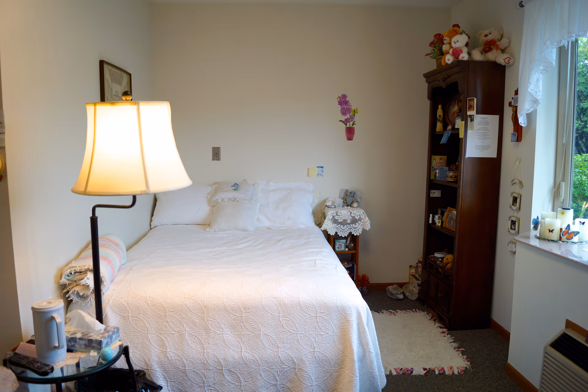 A cozy bedroom in an assisted living facility featuring a neatly made bed with white bedding and pillows. A floor lamp with a white shade stands beside the bed next to a small round glass table holding a tissue box, a remote control, and other small items. On the right side of the bed, there is a wooden shelving unit filled with various personal items and stuffed animals on top. A small table with a lace cover and decorative items sits next to the shelving unit. A window with lace curtains allows natural light to brighten the room, and a small rug is placed on the floor near the shelving unit.