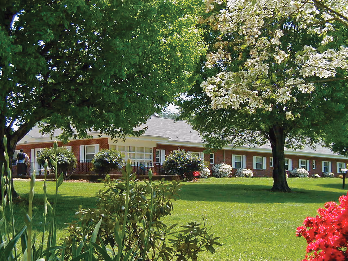Front exterior of a single-story brick assisted living building with a manicured lawn, trees, and flowering shrubs.